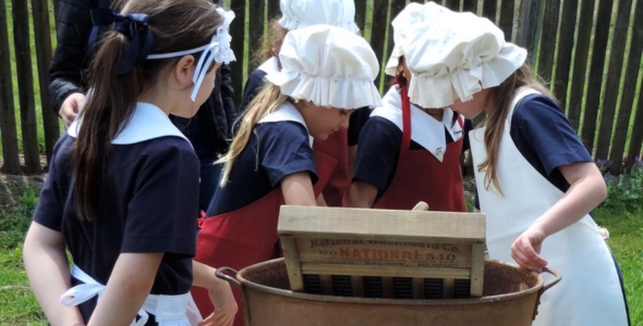 Children in historical costumes use a vintage washboard outdoors.