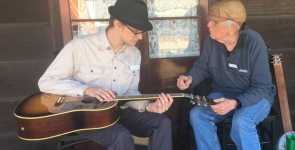 Two men sitting on a porch, one playing a guitar while the other gestures.