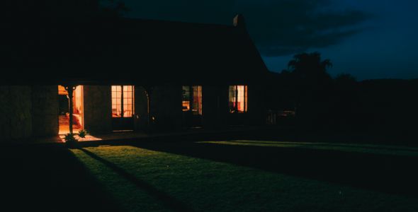 A house with illuminated windows and a dark, grassy yard at night.