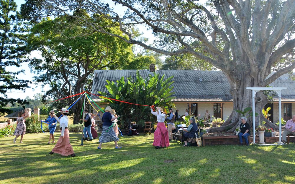 People dancing around a maypole in a garden with a large tree.