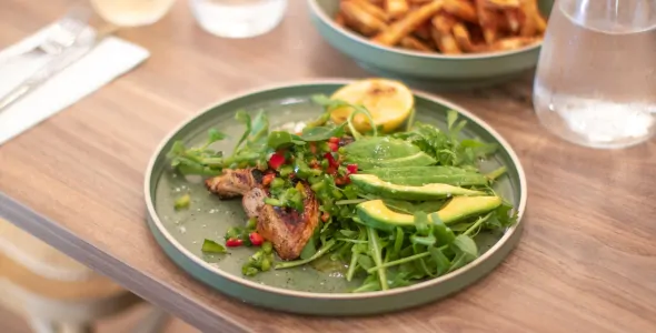 A green plate of a rocket salad with avocado, grilled chicken, pomegranate and a slice of lemon on a wooden table. In the background is a bowl of fries, glass of water and glass of white wine