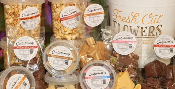A variety of honey products on a table, including honey popcorn, caramel fudge, honeycomb, honeycomb chocolate and a tin in the background with the writing fresh cut flowers on it